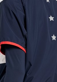 Marineblaues Jackett mit roten Akzenten, kurzen Ärmeln und Mesh-Details. Mit silbernen Sternen entlang der Schultern für ein zusätzliches Designelement. Leichtes Material.