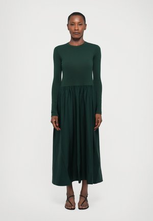 Long-sleeved dark green dress with a fitted bodice and gathered skirt, made from soft fabric. Model wears flat sandals, standing against a plain background.