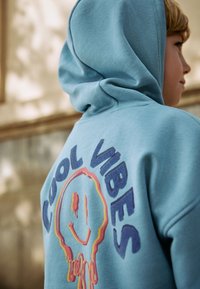 Hellblauer Kapuzenpullover mit dem Text "COOL VIBES" und einer lächelnden Gesichtsgraphik auf der Rückseite, mit tropfenden Akzenten. Weicher, strukturierter Stoff.