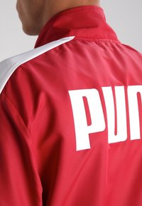 Chaqueta de poliéster roja con acentos blancos, que presenta un gran logo blanco de "PUMA" en la parte posterior. Textura suave y diseño de cuello alto.