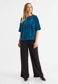 Teal sammetstopp med texturerade vertikala linjer, vida ärmar och rundad halsringning, i kombination med svarta vida byxor och spetsiga svarta skor.
