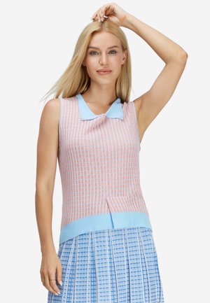 Blonde Frau trägt ein ärmelloses rosa-blau kariertes Oberteil mit blauem Kragen und einen blau gemusterten plissierten Rock, eine Hand zum Kopf erhoben.