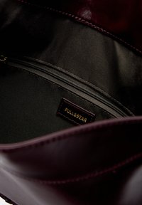 Burgundy leather handbag interior showing a black fabric lining, zipped pocket, and a gold "PULL&BEAR" label. Smooth texture and glossy finish.