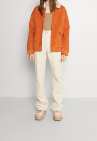 Veste en velours côtelé orange avec un col blanc, haut marron, pantalon large crème et baskets beiges avec des accents roses.