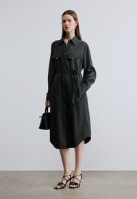 Σκούρο γκρι πουκαμισοφόρεμα με δύο τσέπες στο στήθος, κουμπιά και ζώνη στη μέση. Στυλιζαρισμένο με μαύρα σανδάλια με λουράκια και μικρή μαύρη τσάντα χειρός.