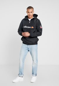 Svart hettejakke med lomme foran; har hvit "ellesse" logo og fargerik detalj. Matcher med lyseblå jeans og hvite joggesko.