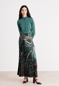 Grüne Bluse mit hohem Ausschnitt, kombiniert mit einem langen, gefältelten Rock mit einem Kettenmuster in erdigen Tönen. Das Model trägt burgunderfarbene Absatzschuhe.
