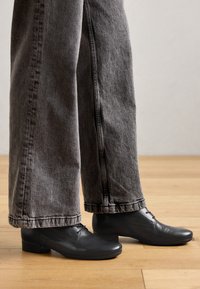 Black leather shoes with a low block heel, rounded toe, and laces, paired with gray wide-leg jeans featuring a raw hem and visible stitching.