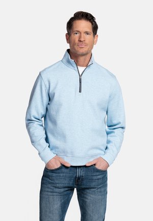 Man draagt een lichtblauwe pullover met ritssluiting tot op de borst en een blauwe spijkerbroek, staand met handen in de zakken tegen een effen witte achtergrond.