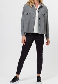 Giacca grigia in tessuto intrecciato con due tasche sul petto, bottoni neri, abbinata a pantaloni neri slim-fit e scarpe da ginnastica nere.
