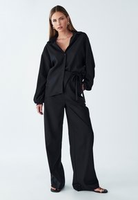 Blusa nera ampia, con colletto e chiusura a bottoni, abbinata a pantaloni neri a gamba larga. Entrambi i capi realizzati in tessuto morbido.