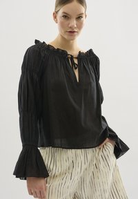 Schwarze, leichte Bluse mit gerafftem Ausschnitt und Bindeband. Mit langen, rüschenbesetzten Ärmeln. Kombiniert mit cremefarbenen, gestreiften, weiten Hosen.