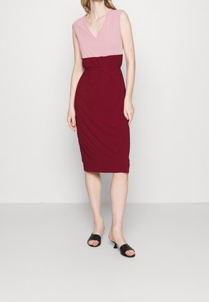 Mujer con vestido sin mangas de bloques de color con parte superior rosa y falda burdeos, de pie sobre un fondo claro y sencillo.
