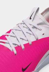 Λευκό και ροζ αθλητικό παπούτσι Nike με λευκά κορδόνια, μαύρο λογότυπο Nike Swoosh στη γλώσσα και στην πλευρά, κοντινή λήψη.