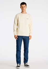 Sudadera crema con cuello redondo, que presenta un pequeño logo negro. Combinada con jeans azules y zapatos negros. El modelo se encuentra de pie contra un fondo blanco.