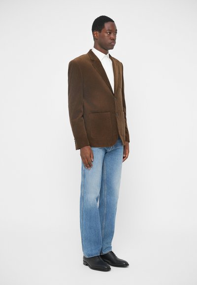 Brun fløjlsblazer med to frontlommer, parret med lyse lige ben jeans og sorte sko, på en neutral baggrund.