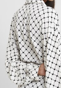 Blusa a maniche lunghe in un tessuto leggero, con un motivo di cuori neri e catene su uno sfondo bianco. Dettaglio delle maniche arricciate visibile.