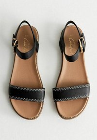 Sandalias de cuero negro con una banda ancha, correa ajustable en el tobillo y plantilla en tono marrón. Un pespunte blanco añade detalle a lo largo de los bordes de la banda.