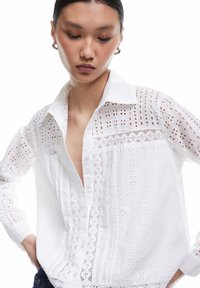 Ung kvinde med mørkt hår iført en hvid øjelet blondebluse og sølv hoop-øreringe, ser ned med hænderne på hoften.