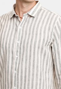 Camicia leggera in lino color crema con righe verticali grigie. Presenta un colletto button-down e una pattina anteriore con bottoni bianchi.