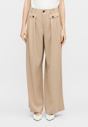 Žena v béžových širokých nohaviciach s vreckami so zapínaním na gombíky a bielych topánkach na špic s čipkovými detailmi, stojaca na bielom pozadí.