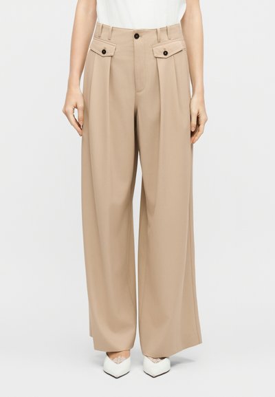 Femme portant un pantalon beige à jambes larges avec des poches à rabat boutonnées et des chaussures blanches à bout pointu avec des accents en dentelle, debout devant un fond blanc.