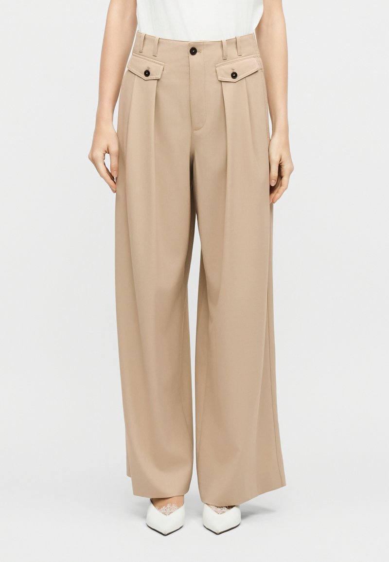 Γυναίκα που φοράει μπεζ παντελόνι με φαρδιά πόδια και τσέπες με πατιλέτα και κουμπιά, μαζί με λευκά παπούτσια μυτερά με λεπτομέρειες από κορδόνια, στέκεται μπροστά σε λευκό φόντο.