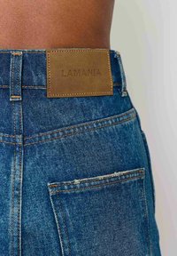 Džínsy v stredne modrej farbe s hnedou koženou nášivkou na zadnej strane s nápisom "LAMANIA", so viditeľným prešívaním a detailmi vreciek.