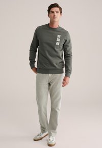 Joven con un sudadera verde con diseños de hojas blancas, jeans gris claro y zapatillas blancas, de pie contra un fondo liso.