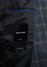 Schwarzes Satin-Innenfutter mit einem Etikett, das "JACK & JONES" liest. Sichtbare Größenetiketten: UK 38R, 48. Blau gemusterter Stoff auf der rechten Seite.