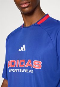 Sininen urheilupaita punaisilla yksityiskohdilla, lyhyet hihat ja pyöreä kaula-aukko. Varustettu valkoisella Adidas-logolla ja suurella punaisella tekstillä, jossa lukee "ADIDAS SPORTSWEAR."