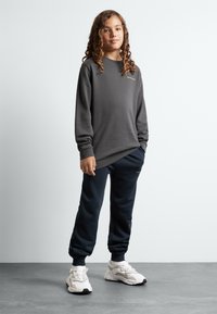 Sweat à manches longues gris avec des poignets côtelés, associé à un pantalon de jogging marine. Des chaussures de sport blanches complètent la tenue. Design simple, coupe confortable.