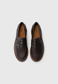 Mocassins en cuir marron avec un bout rond, une sangle décorative et des coutures détaillées. Doublure intérieure douce et semelle texturée pour une bonne adhérence.