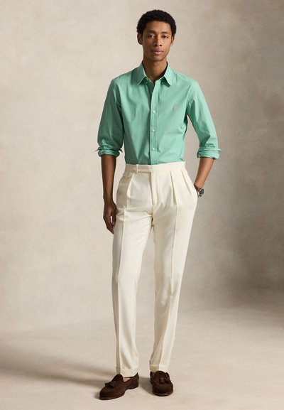 Mand står med hænderne i lommerne iført en opsmøgede grøn skjorte med knapper, cremefarvede bukser med høj talje og brune loafers med frynser mod en neutral baggrund.
