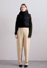Svart puffervest over en grønn genser, beige bukser med elastisk liv, og svarte spisse hæler. Glatte teksturer, enkle designelementer.