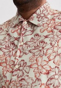 Chemise beige à motif floral rouge et rose foncé, avec un col standard, une fermeture à boutons à l'avant, et un tissu texturé.