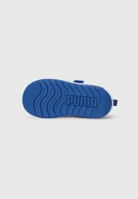 Sandale blanche d'enfant avec semelle en caoutchouc bleu montrant un motif de rainures ondulées et le logo "PUMA" au dos, photographiée sur fond blanc.