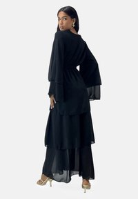 Zwarte gelaagde jurk met wijde mouwen, gemaakt van lichtgewicht stof. Heeft een getailleerde taille en een vloeiende zoom tot aan de enkels.