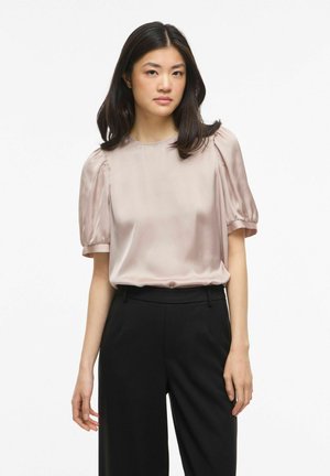 Ung kvinde med glat sort hår iført en lyserød satinbluse med ballonærmer og sorte bukser med høj talje, stående foran en hvid baggrund.