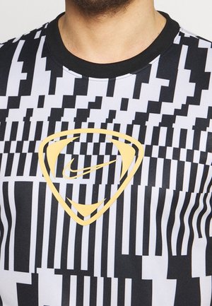 Detail osoby nosící černobíle geometricky vzorovanou košili se žlutým štítem a logem Nike swoosh na hrudi.