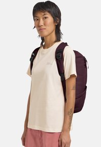 Giovane con capelli corti e scuri che indossa una t-shirt crema, pantaloni rosa e uno zaino viola scuro, in piedi di lato su uno sfondo bianco.