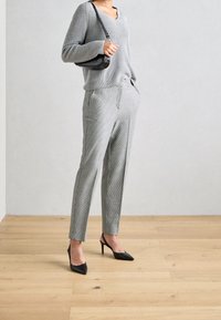Femme portant un pull en tricot gris et un pantalon tailleur gris, tenant un sac à main noir, se tenant sur un sol en bois clair contre un mur uni.
