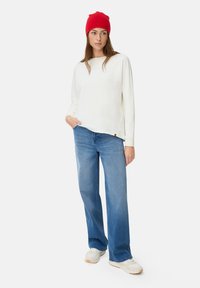 Vrouw in een rode beanie, witte langärmige top, blauwe wijde jeans en witte sneakers, staand met één hand in de zak.