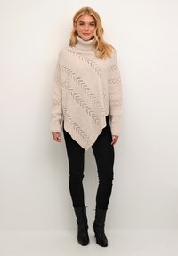 Beige pletený pončo s vysokým límcem, s cik-cak vzory a žebrovanou strukturou. Kombinováno s černými slim kalhotami a kotníkovými botami.