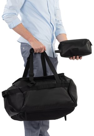 Personne portant une chemise bleu clair et un pantalon bleu, tenant un grand sac de sport noir dans une main et une petite trousse de toilette noire dans l'autre.