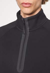 Sudadera de forro polar negra con un cuello alto, que presenta una cremallera de un cuarto y un borde de cremallera suave y en contraste. Tela suave y elástica.