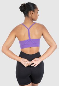 Violetti urheiluliivi, jossa on ohut hihna ja ribbestehoste, yhdistettynä korkeavyötäröisiin mustiin shortseihin. Takaa näkyy ristiselkädesign.