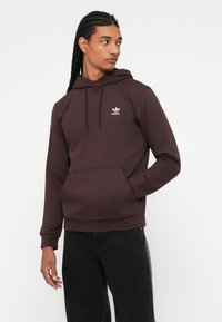 Jaunas vyras su pintomis plaukai vilki tamsiai purpurinę Adidas džemperį ir juodus džinsus, žiūri į kairę, laikydamas vieną ranką kišenėje.