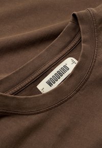 Κοντινό πλάνο του γιακά ενός καφέ t-shirt με στρογγυλή λαιμόκοψη, με εμφανή λευκή ετικέτα που δείχνει το όνομα της μάρκας "WOODBIRD" και λεπτομέρειες μεγέθους.