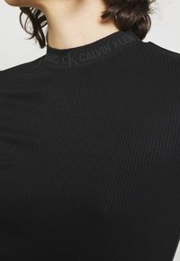 Czarna top z prążkowanej tkaniny o wysokim dekolcie. Posiada subtelne logo Calvin Klein wzdłuż kołnierza. Gładka tekstura i dopasowany kształt.